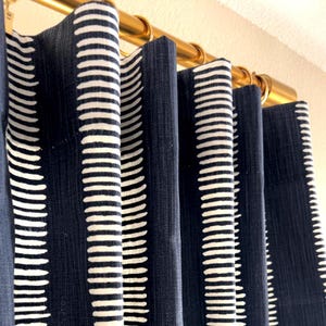 Puede incluir: Primer plano de cortinas azul marino con un patrón repetitivo de rayas blancas verticales. Las cortinas cuelgan de una barra dorada. La tela tiene una apariencia texturizada, agregando profundidad al diseño. La estética general es moderna y elegante.