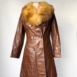 May include: A brown leather coat with a faux fur collar. The double-breasted coat features large, round buttons and a tailored fit. The coat is displayed on a mannequin against a white background, showcasing its vintage style.