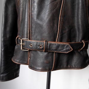 May include: Close-up of a vintage brown leather jacket with a belt. The jacket has a distressed appearance with visible wear and tear. The belt features a metal buckle and is cinched around the waist. The jacket's seams are highlighted with lighter brown stitching.