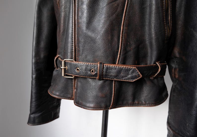 May include: Close-up of a vintage brown leather jacket with a belt. The jacket has a distressed appearance with visible wear and tear. The belt features a metal buckle and is cinched around the waist. The jacket's seams are highlighted with lighter brown stitching.