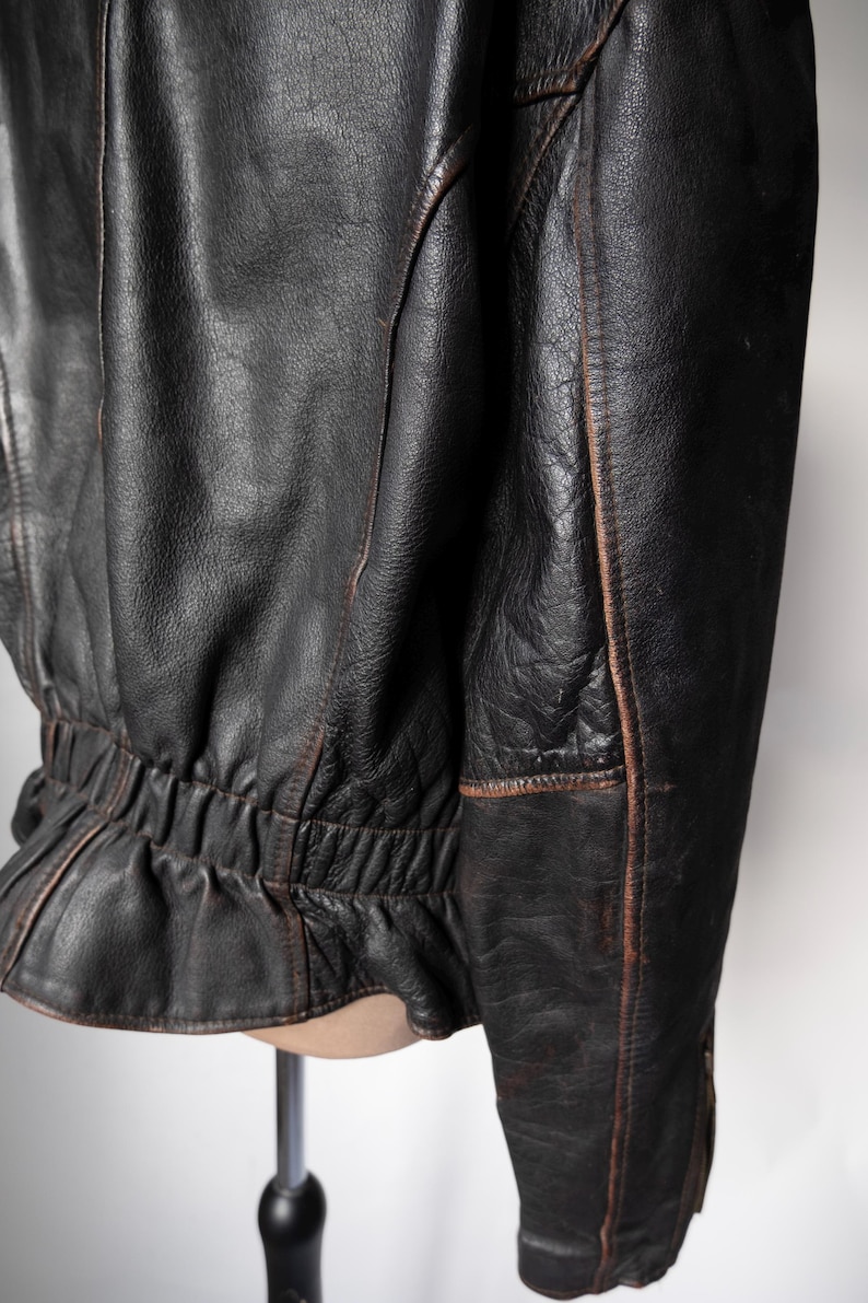 May include: Close-up of a black leather jacket with visible wear and tear. The jacket has a cinched waist and detailed stitching along the seams. The leather appears distressed with brown accents.