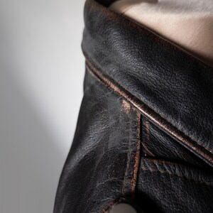 May include: Close-up of a black leather jacket with a distressed, vintage look. The jacket features a notched collar, visible stitching, and a silver snap closure. The leather shows signs of wear, with a brown trim.