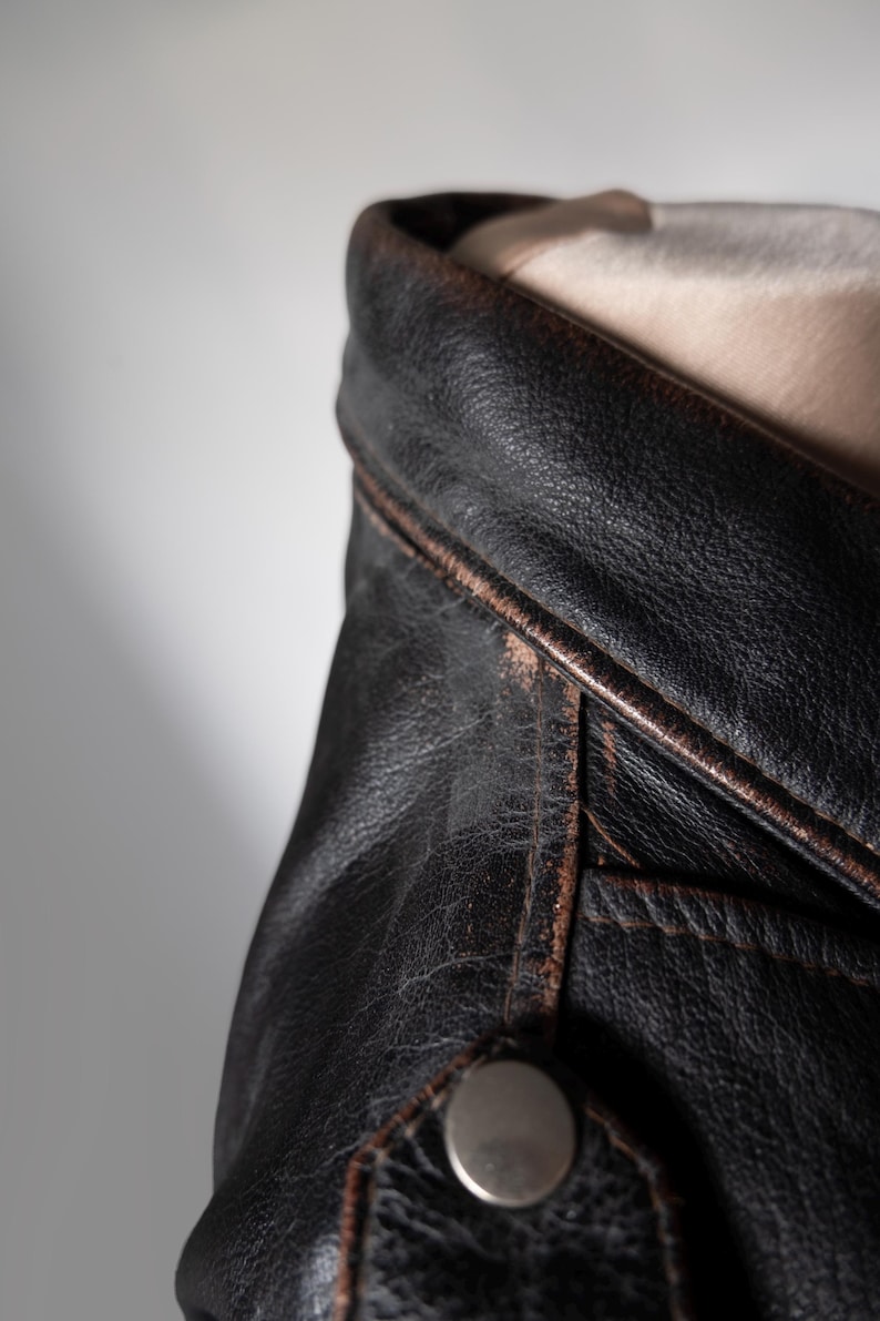 May include: Close-up of a black leather jacket with a distressed, vintage look. The jacket features a notched collar, visible stitching, and a silver snap closure. The leather shows signs of wear, with a brown trim.