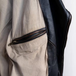 May include: Close-up of a black leather jacket interior. The jacket features a tan lining with a zippered pocket. The edges of the jacket are black leather with visible stitching. The zipper is a dark bronze color.