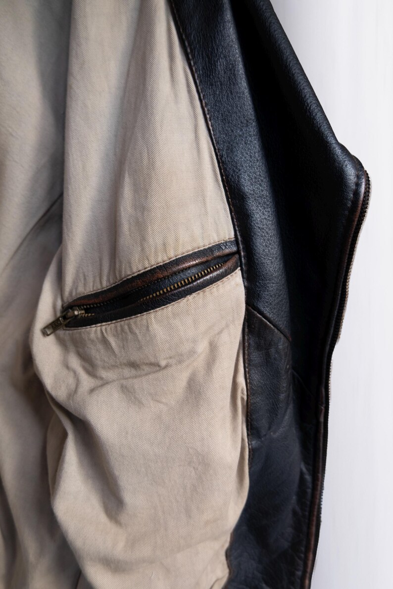 May include: Close-up of a black leather jacket interior. The jacket features a tan lining with a zippered pocket. The edges of the jacket are black leather with visible stitching. The zipper is a dark bronze color.