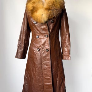 May include: A brown leather coat with a fur collar. The coat has a double-breasted design with four buttons and a long length. The fur collar is a mix of brown and tan. The coat is displayed on a mannequin.