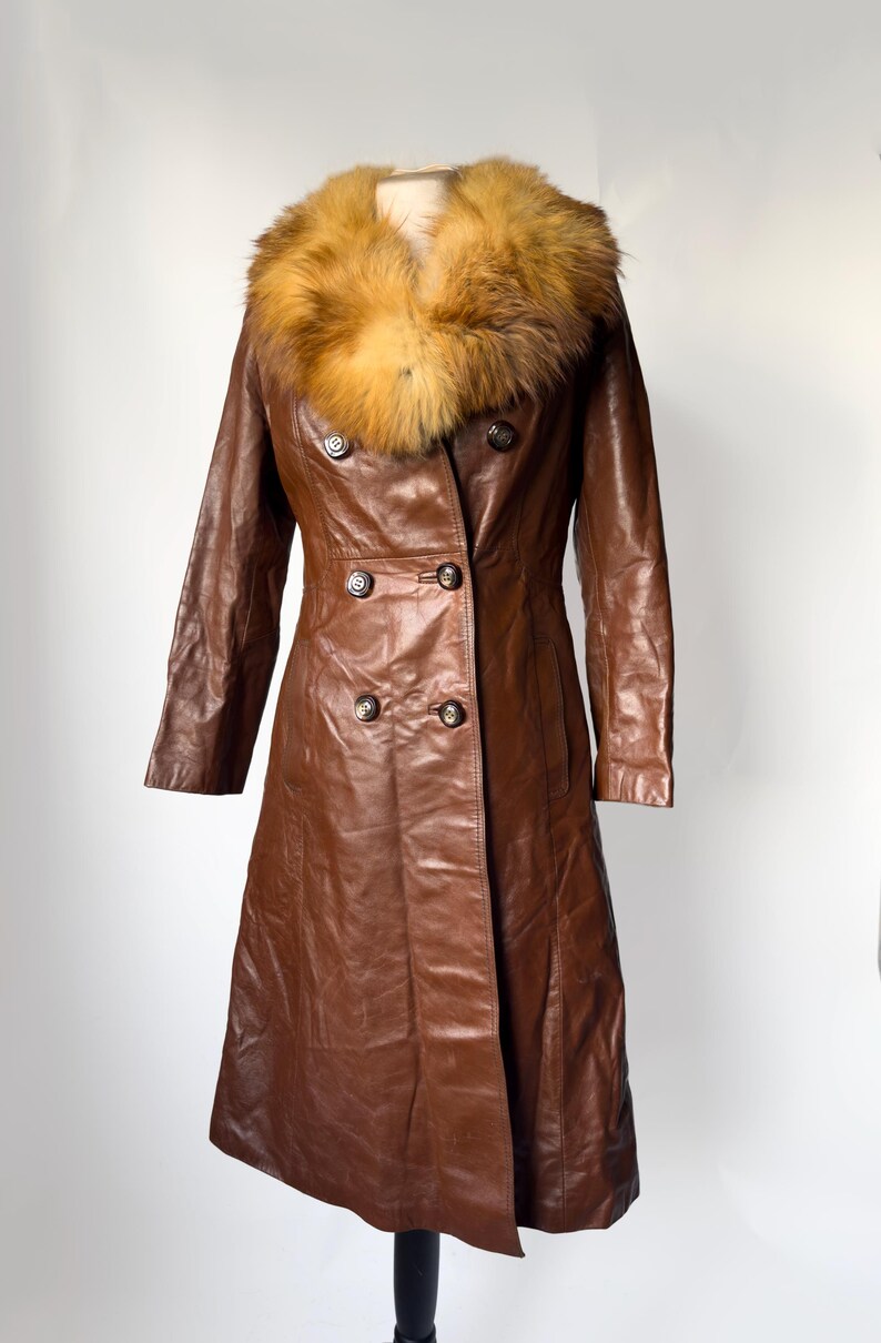 May include: A brown leather coat with a fur collar. The coat has a double-breasted design with four buttons and a long length. The fur collar is a mix of brown and tan. The coat is displayed on a mannequin.