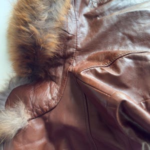 May include: Close-up of a brown leather jacket with a fur-lined hood. The jacket has a textured appearance and visible stitching details. The fur trim is a mix of brown and gray tones, adding a luxurious touch to the outerwear.