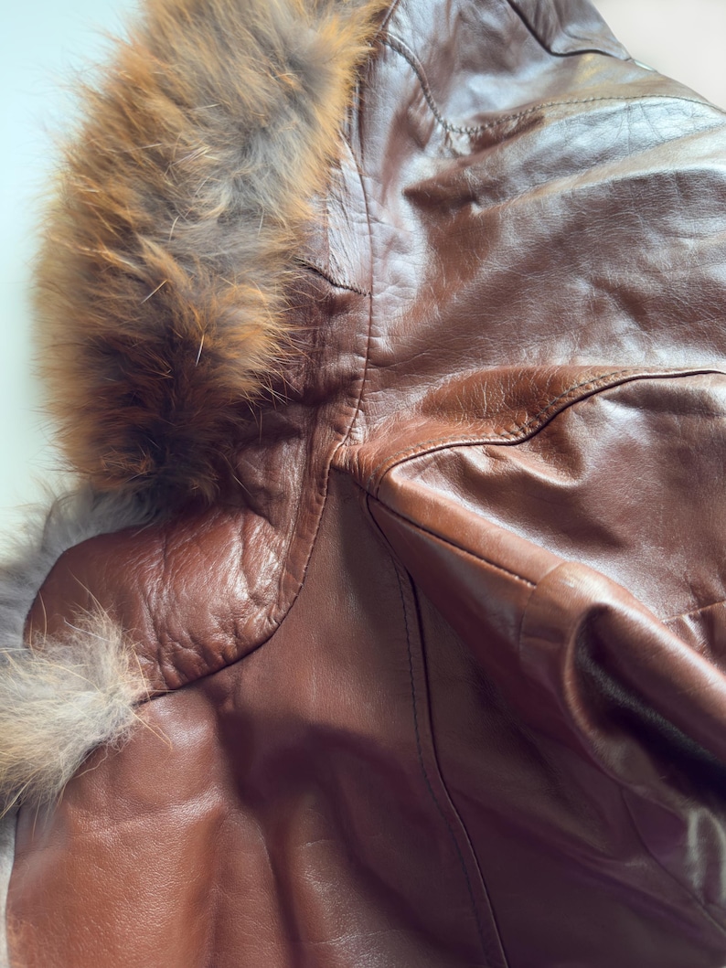 May include: Close-up of a brown leather jacket with a fur-lined hood. The jacket has a textured appearance and visible stitching details. The fur trim is a mix of brown and gray tones, adding a luxurious touch to the outerwear.
