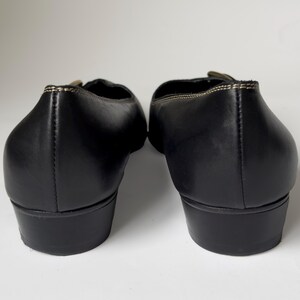 May include: A pair of black leather heeled shoes with a gold buckle detail. The shoes have a pointed toe and a low heel.