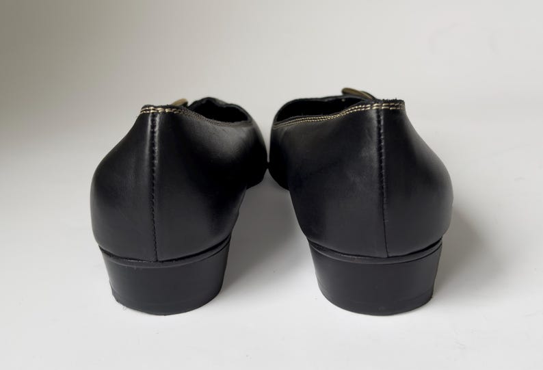May include: A pair of black leather heeled shoes with a gold buckle detail. The shoes have a pointed toe and a low heel.