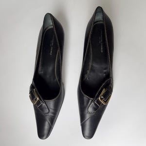 May include: A pair of black leather heeled shoes with a pointed toe and a gold buckle. The shoes have a stitched detail around the edges.