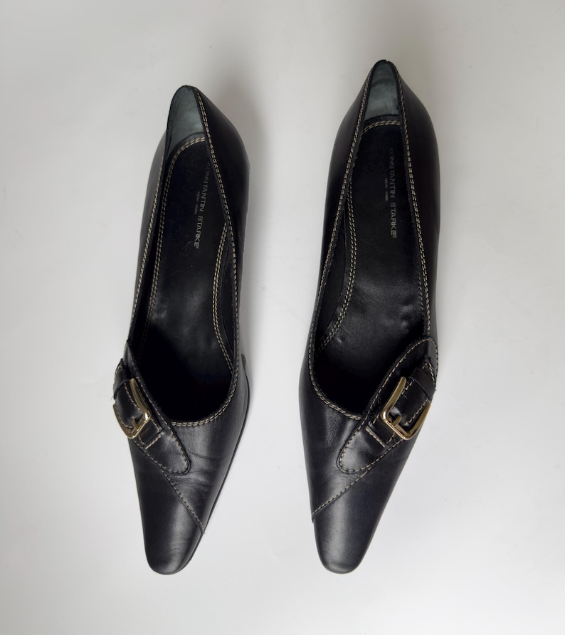 May include: A pair of black leather heeled shoes with a pointed toe and a gold buckle. The shoes have a stitched detail around the edges.