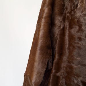 May include: A close-up of a brown fur coat sleeve. The coat has a rich, dark brown color and a soft, textured appearance. The fur appears dense and luxurious, suggesting warmth and quality. The background is a plain white.
