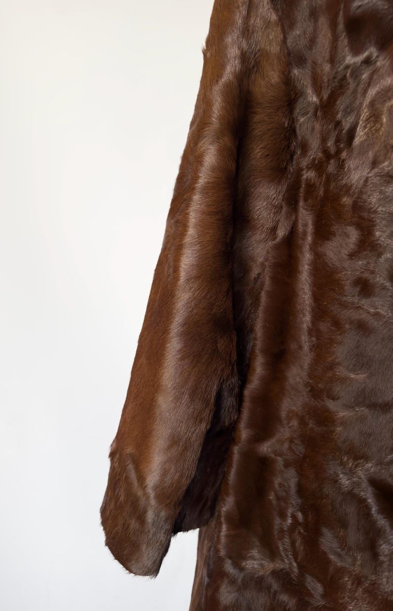 May include: A close-up of a brown fur coat sleeve. The coat has a rich, dark brown color and a soft, textured appearance. The fur appears dense and luxurious, suggesting warmth and quality. The background is a plain white.