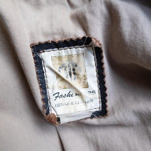 May include: Close-up of a leather jacket's interior, showcasing a beige lining and a rectangular label. The label, framed in black with a scalloped edge, reads "Fashion" and "GENUINE LEATHER".