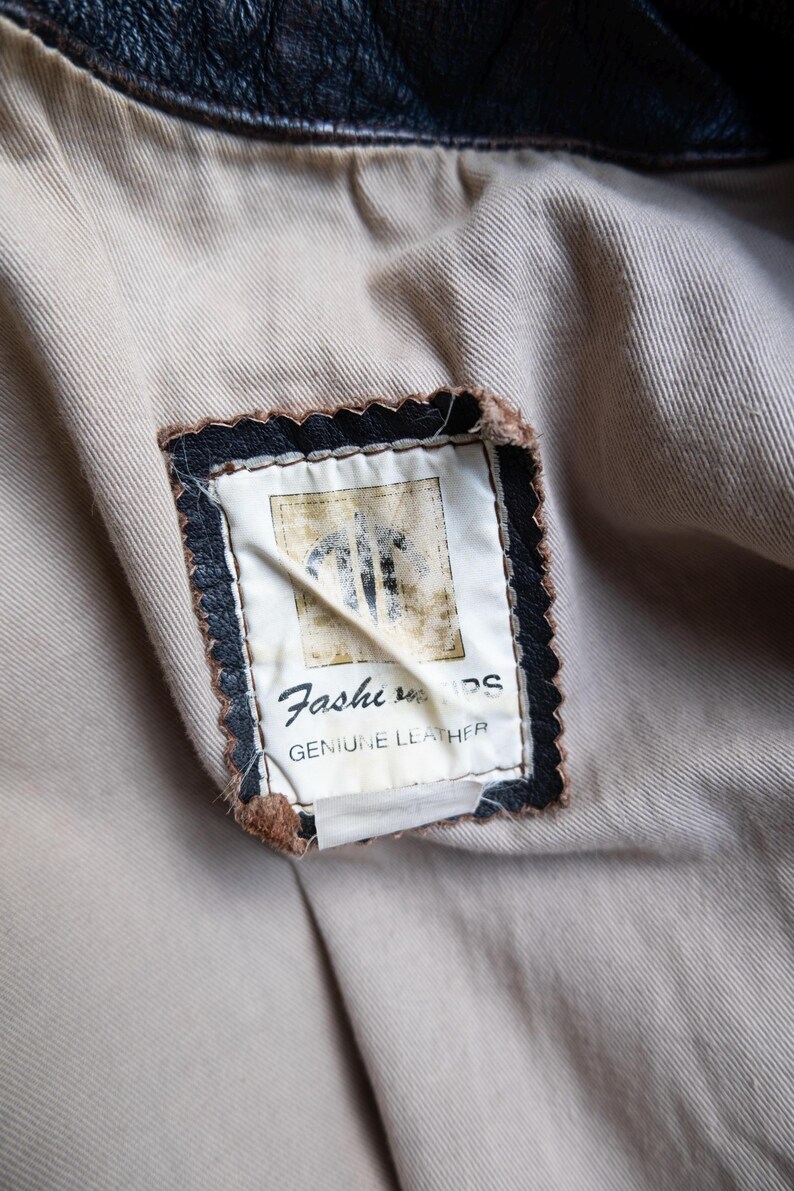 May include: Close-up of a leather jacket's interior, showcasing a beige lining and a rectangular label. The label, framed in black with a scalloped edge, reads "Fashion" and "GENUINE LEATHER".