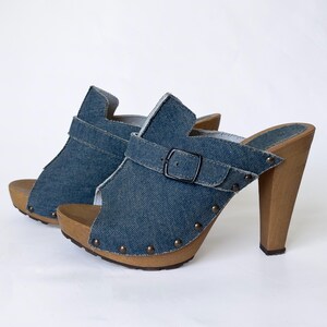 May include: A pair of denim clogs with wooden heels. The open-toe shoes feature a blue denim upper with a buckle strap and small metal studs. The heels are a light brown color.