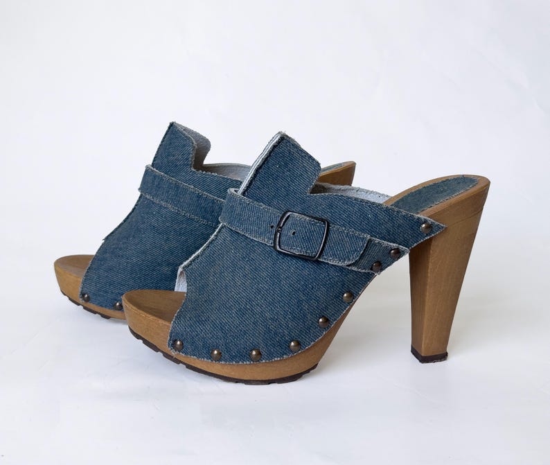 May include: A pair of denim clogs with wooden heels. The open-toe shoes feature a blue denim upper with a buckle strap and small metal studs. The heels are a light brown color.