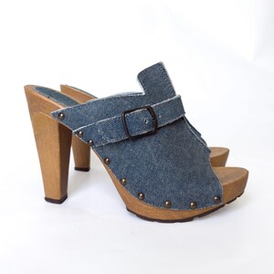 May include: Denim clogs with wooden heels and soles. The open-toe shoes feature a blue denim upper with a buckle strap and small metal studs. The heels are a light brown color.