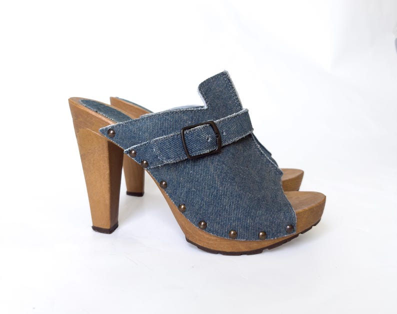 May include: Denim clogs with wooden heels and soles. The open-toe shoes feature a blue denim upper with a buckle strap and small metal studs. The heels are a light brown color.