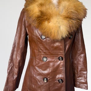 May include: A brown leather coat with a fur collar. The double-breasted coat has dark buttons and a tailored fit. The fur collar is a light brown color. The coat is displayed on a mannequin.