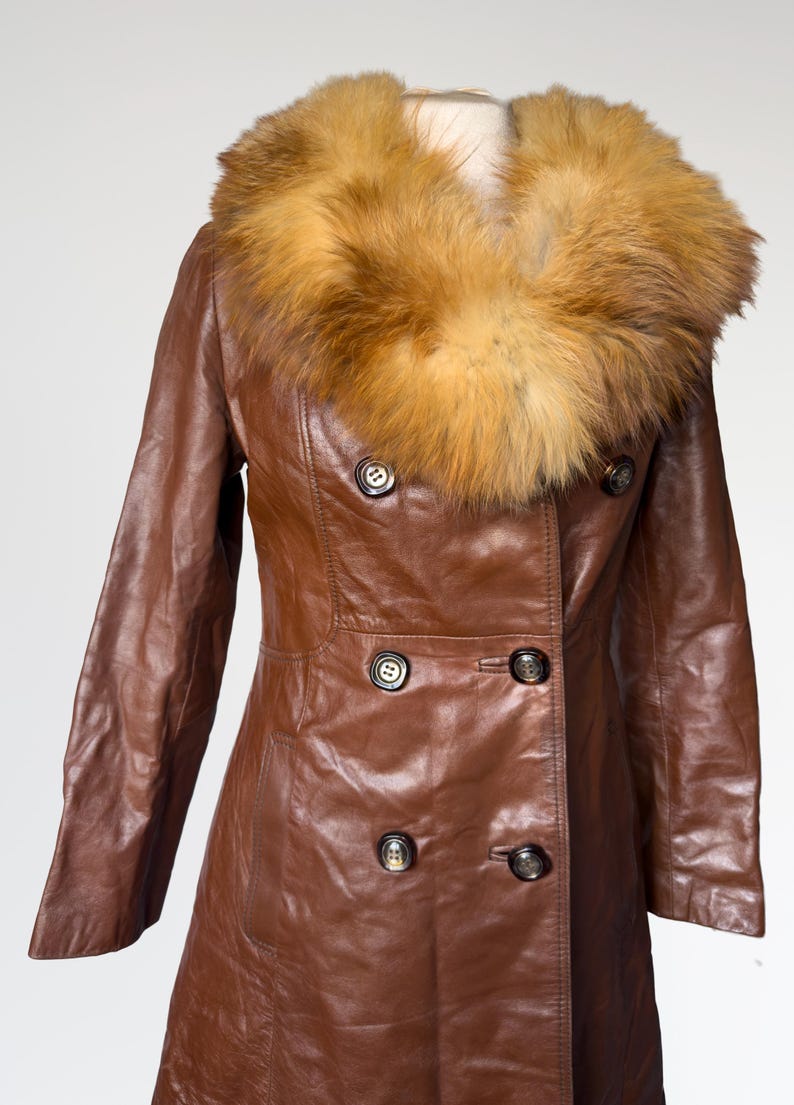 May include: A brown leather coat with a fur collar. The double-breasted coat has dark buttons and a tailored fit. The fur collar is a light brown color. The coat is displayed on a mannequin.