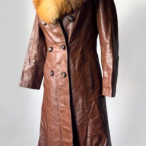 May include: A brown leather coat with a fur collar. The coat has a double-breasted design with four buttons and two pockets. The fur collar is a mix of brown and tan colors. The coat is displayed on a black stand.