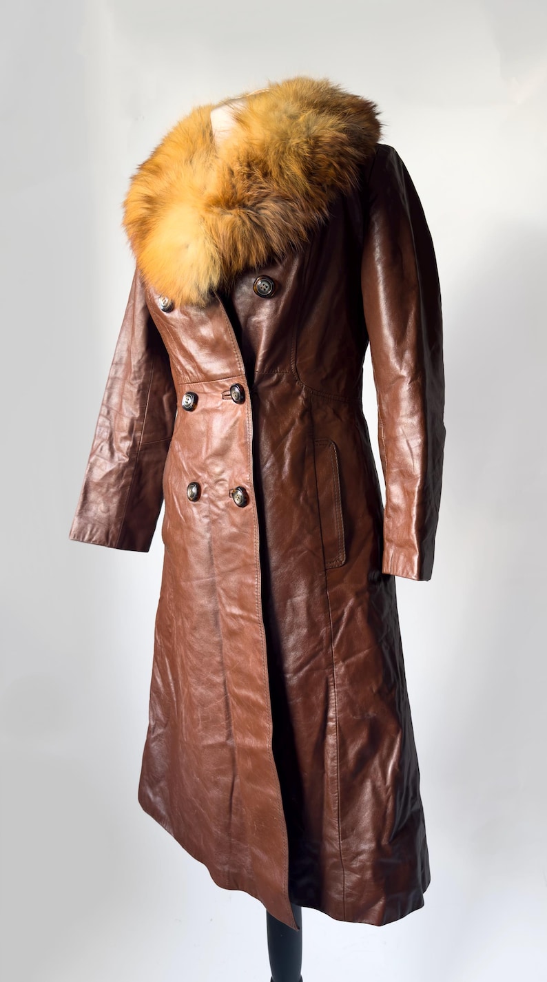 May include: A brown leather coat with a fur collar. The coat has a double-breasted design with four buttons and two pockets. The fur collar is a mix of brown and tan colors. The coat is displayed on a black stand.