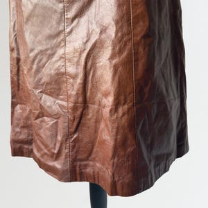 May include: A brown leather skirt with vertical seams. The skirt is displayed on a black mannequin stand against a white background. The leather has a textured appearance.