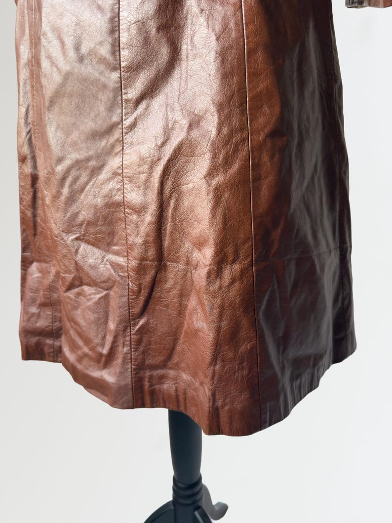 May include: A brown leather skirt with vertical seams. The skirt is displayed on a black mannequin stand against a white background. The leather has a textured appearance.