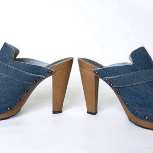 May include: A pair of denim high-heeled clogs. The open-toe shoes have a blue denim upper with a strap and wooden heels. The soles are also made of wood and are studded with small metal rivets.