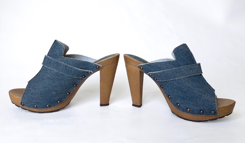 May include: A pair of denim high-heeled clogs. The open-toe shoes have a blue denim upper with a strap and wooden heels. The soles are also made of wood and are studded with small metal rivets.