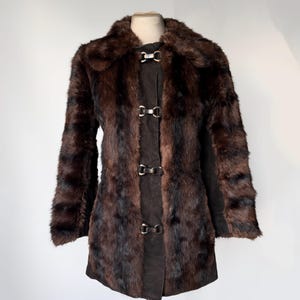 以下が含まれることがあります： ノッチカラーと長袖の茶色のフェイクファーコート。 コートは、3つの装飾的な金属製の留め金が付いた、前面に濃い茶色のスエードパネルを備えています。 コートはマネキンに展示されています。