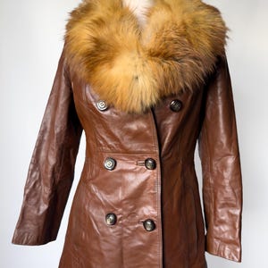 May include: A brown leather coat with a large, fluffy, tan and brown fur collar. The coat has a double-breasted design with dark buttons and a tailored fit. The coat is displayed on a mannequin against a white background.