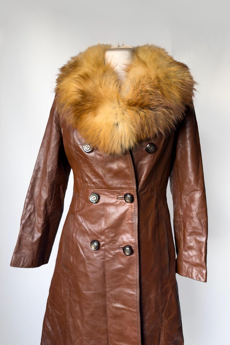 May include: A brown leather coat with a large, fluffy, tan and brown fur collar. The coat has a double-breasted design with dark buttons and a tailored fit. The coat is displayed on a mannequin against a white background.