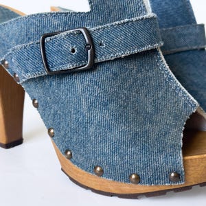 May include: Blue denim clogs with a peep-toe design. The shoes feature a wooden sole, a buckle closure, and metal stud accents. The high heel is made of wood.