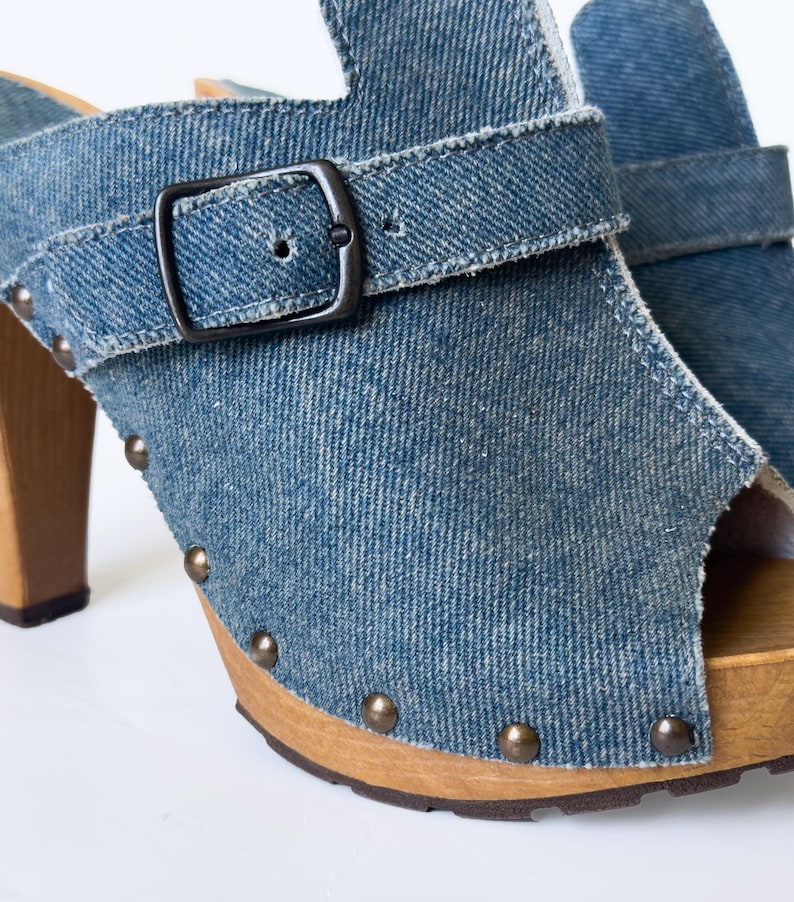 May include: Blue denim clogs with a peep-toe design. The shoes feature a wooden sole, a buckle closure, and metal stud accents. The high heel is made of wood.