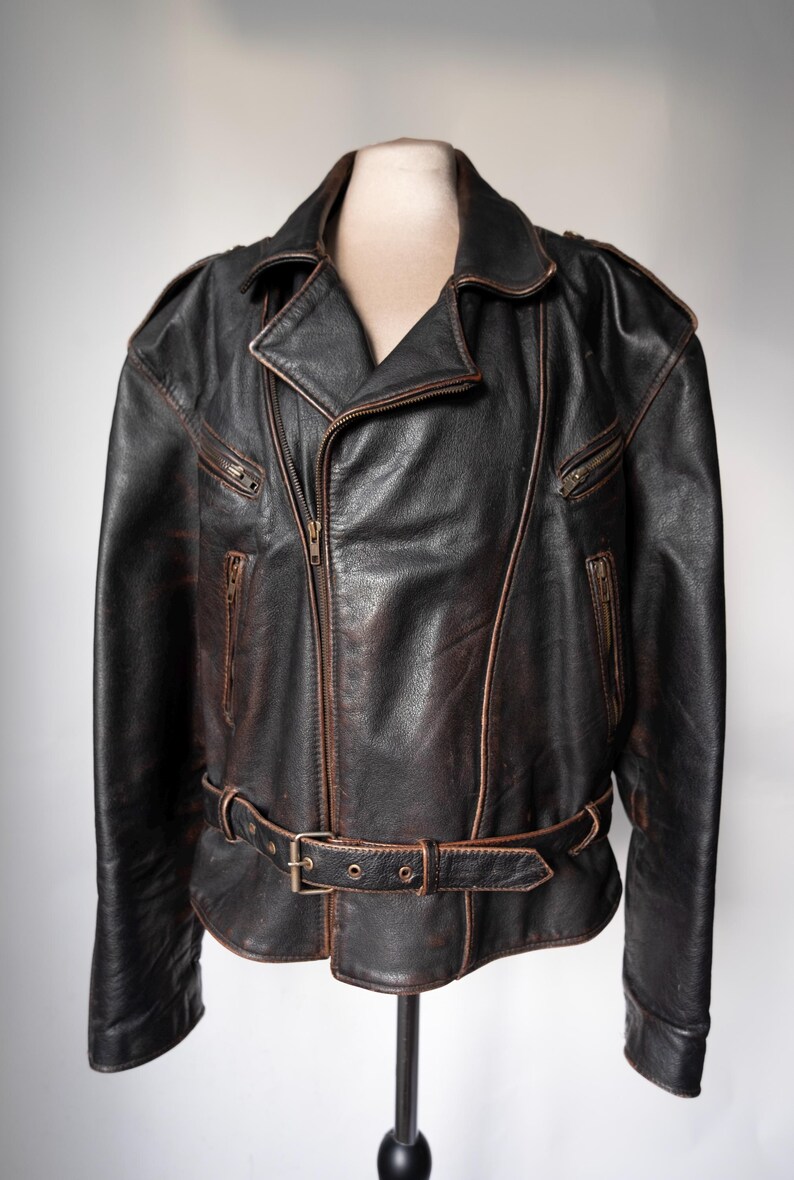 May include: A black leather jacket with a brown distressed finish. The jacket features a notched collar, a front zipper closure, and multiple zippered pockets. It has a belt at the waist and is displayed on a mannequin.