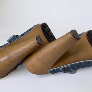 May include: Wooden-heeled clogs with denim straps. The clogs have a light brown wooden base and a dark brown sole. The denim straps are a faded blue and are secured with small silver studs. The heels are approximately 3 inches tall.