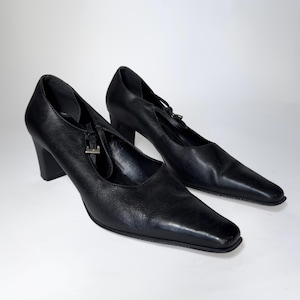 Puede incluir: Un par de zapatos de tacón de cuero negro con punta cuadrada y una correa en la parte superior. Los tacones miden aproximadamente 7.6 cm de alto. Los zapatos están sobre un fondo blanco.