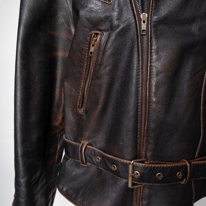 May include: Close-up of a distressed brown leather jacket with a zipper closure and multiple pockets. The jacket features a belt at the waist and a worn, vintage appearance. The leather has a textured, aged look.