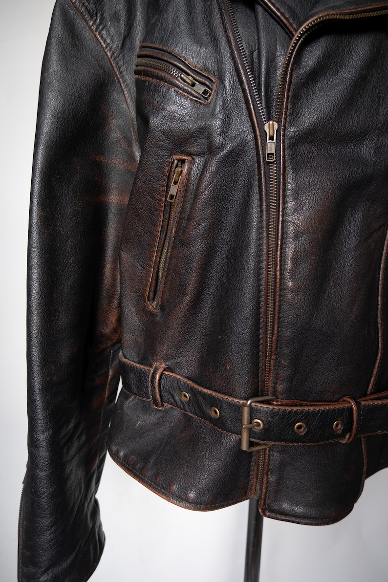 May include: Close-up of a distressed brown leather jacket with a zipper closure and multiple pockets. The jacket features a belt at the waist and a worn, vintage appearance. The leather has a textured, aged look.