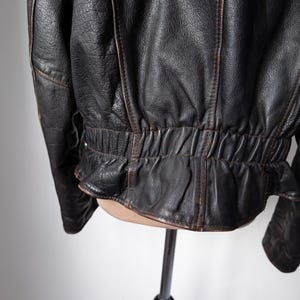 May include: Close-up of a black leather jacket with brown accents. The jacket has a textured surface and a gathered waist detail. The stitching is visible, and the leather appears worn.
