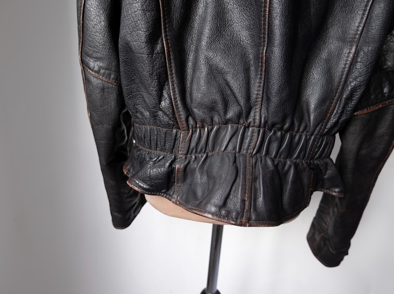 May include: Close-up of a black leather jacket with brown accents. The jacket has a textured surface and a gathered waist detail. The stitching is visible, and the leather appears worn.