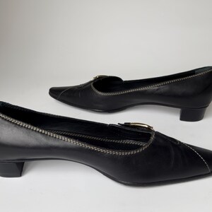 May include: A pair of black leather heeled shoes with a pointed toe and a buckle detail. The shoes have a low heel and a white stitching detail.