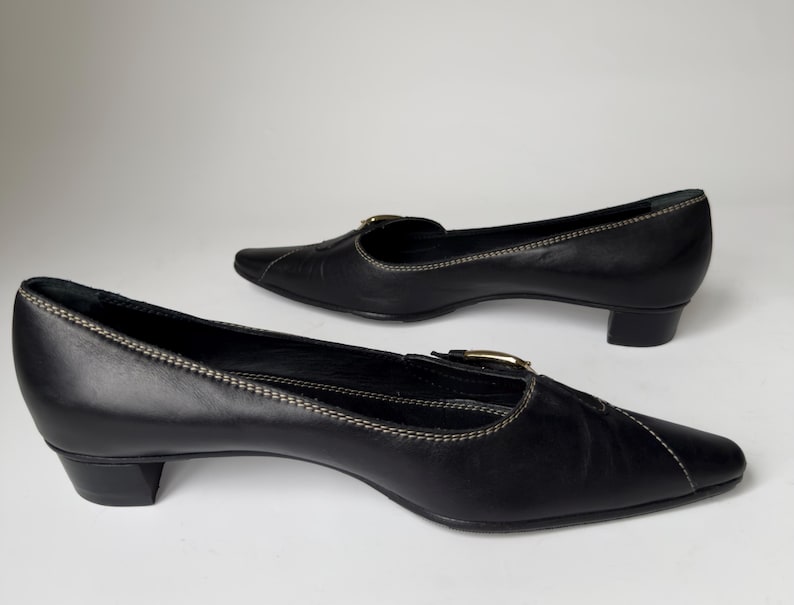 May include: A pair of black leather heeled shoes with a pointed toe and a buckle detail. The shoes have a low heel and a white stitching detail.