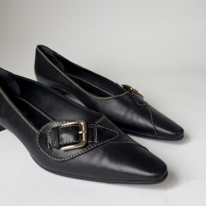 May include: Pair of black leather heeled shoes with a pointed toe and a buckle detail. The shoes have a subtle stitch detail around the edges.