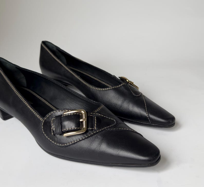 May include: Pair of black leather heeled shoes with a pointed toe and a buckle detail. The shoes have a subtle stitch detail around the edges.