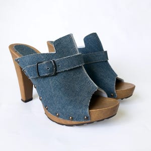 May include: A pair of denim clogs with wooden heels and soles. The blue denim uppers have a buckle strap and open-toe design. The heels are approximately 10 cm tall, and the soles are studded with small metal rivets.